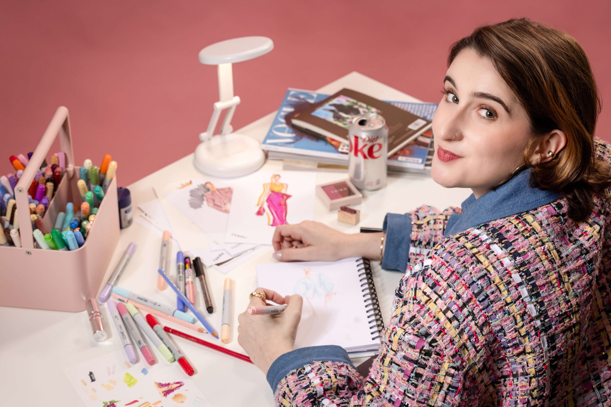 woman in pink and blue jacket drawing fashion illustrations for live events at a white desk
