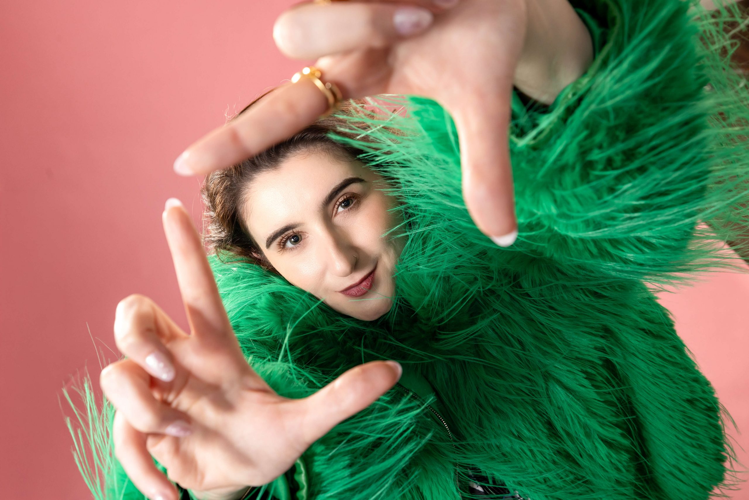 woman in green furry coat holding up hands like a camera and smiling against a pink background